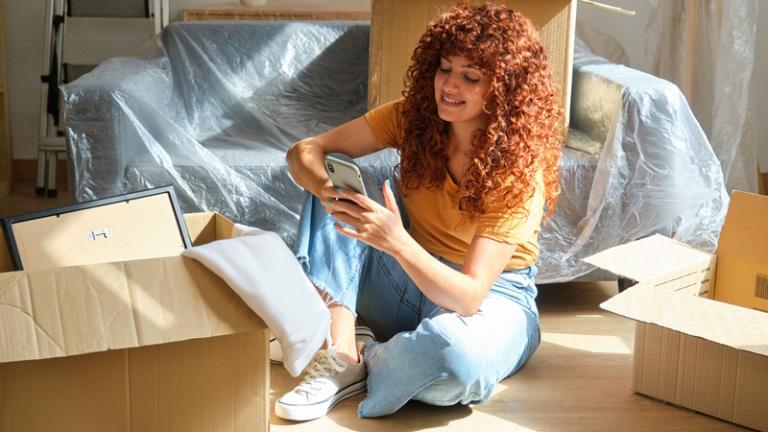 Woman sits among moving boxes looking at her phone